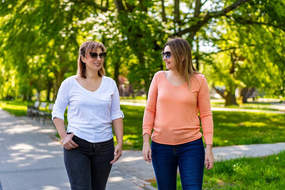 Two women walking together and smiling at each other in a leafy park.