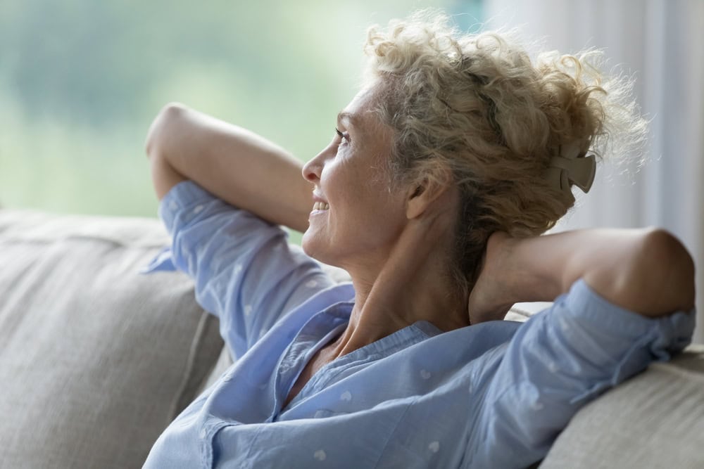 “Woman relaxing on a sofa with her hands behind her head, looking towards a window.”